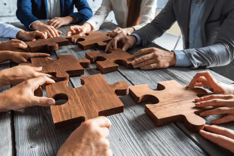 business-people-team-sitting-around-meeting-table-assembling-wooden-jigsaw-puzzle-pieces-unity-cooperation-ideas-concept_394555-647 (1)